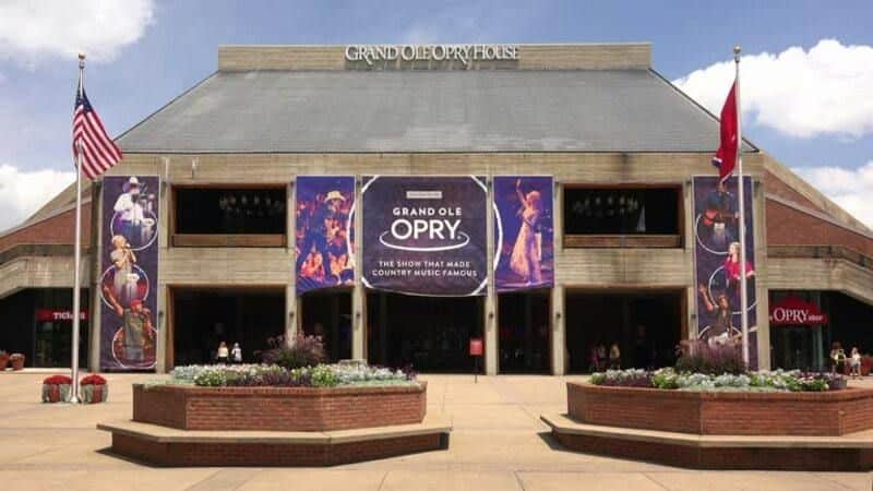 The Grand Ole Opry House with banners and flags on display, a few people near the entrance, and planters with flowers in the foreground.