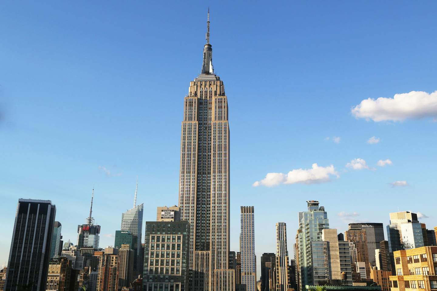 Empire State Building City Cards & Passes (New York) Tall skyscraper with a spire, surrounded by other high-rise buildings under a clear blue sky with a few clouds.