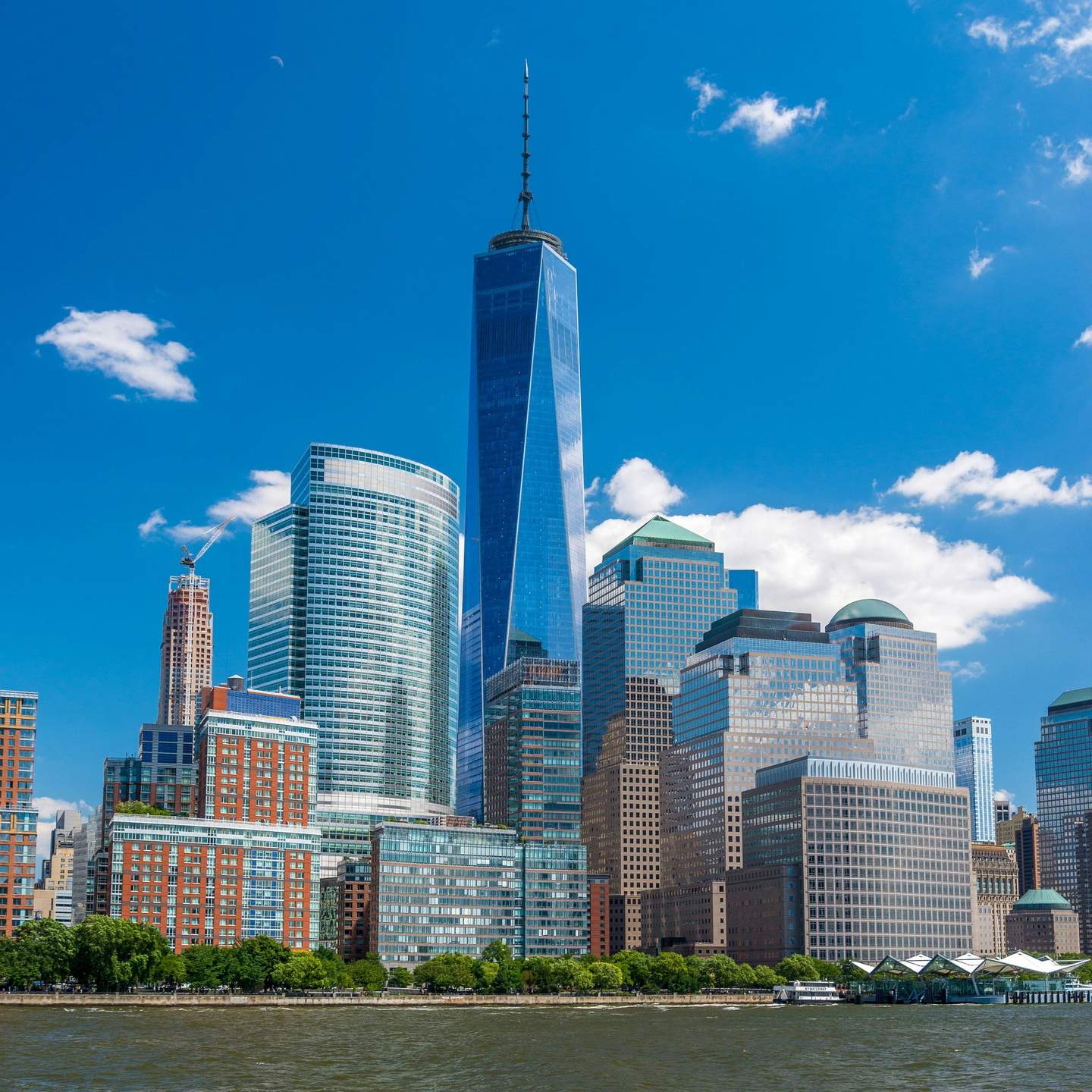 One World Observatory City Cards & Passes (New York) Several modern skyscrapers, including a tall, pointed tower, stand along the waterfront under a clear blue sky.