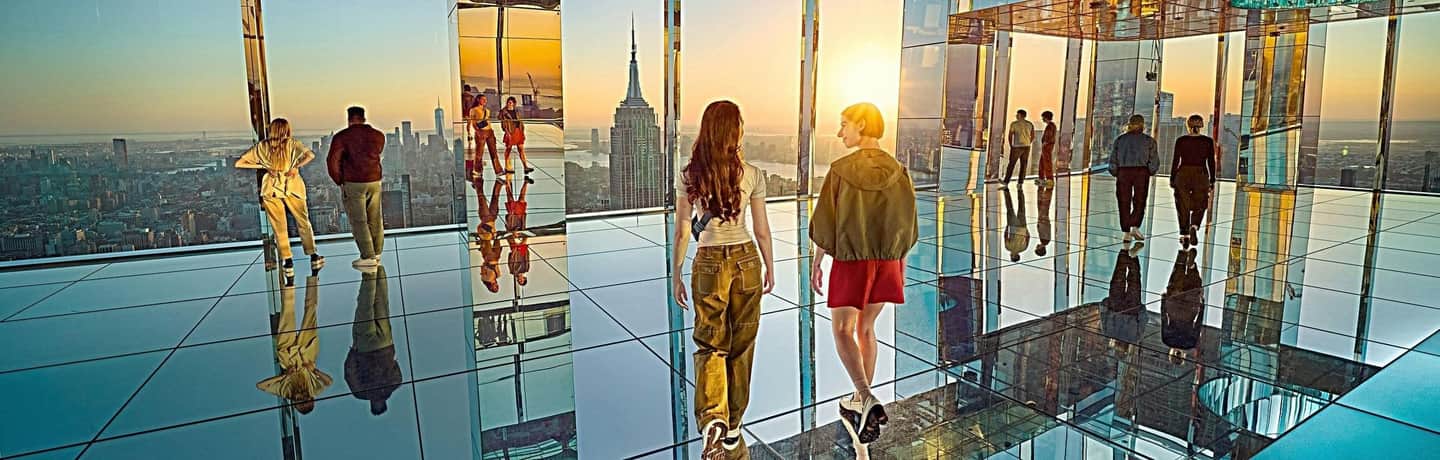 People stand and walk on a mirrored observation deck with a view of a city skyline at sunset.