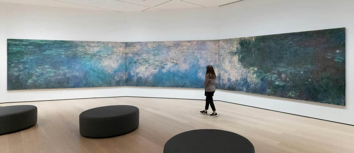 Une personne regarde une grande peinture murale de nénuphars dans une galerie minimaliste avec des sièges gris foncé.