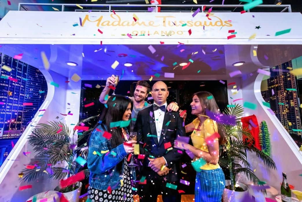 Three people celebrate with confetti and drinks around a wax figure in a tuxedo at Madame Tussauds Orlando.