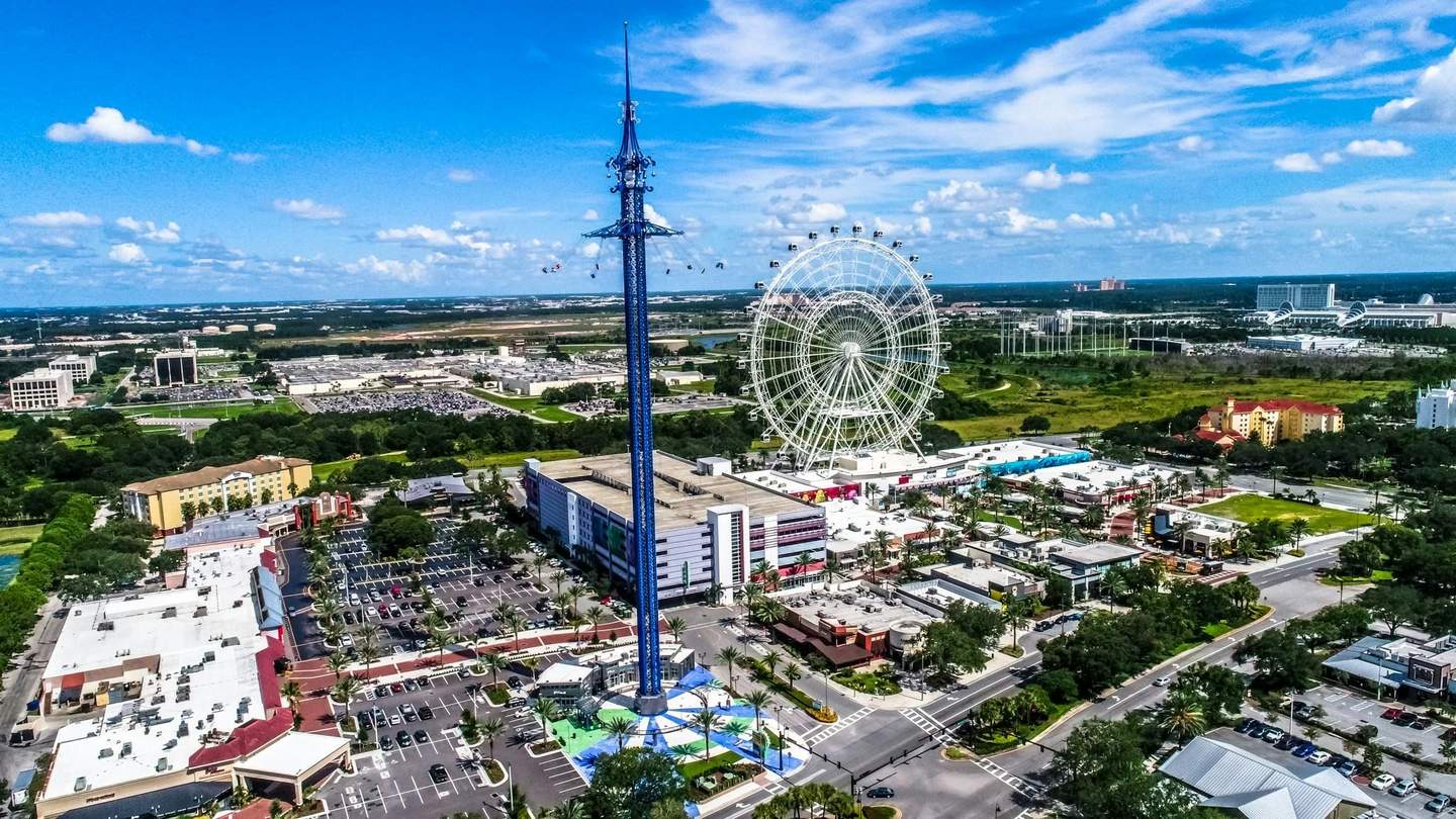 The Orlando Eye City Card (Orlando) Aerial view of an amusement park with a tall swing ride, Ferris wheel, parking lots, buildings, and surrounding greenery under a blue sky.