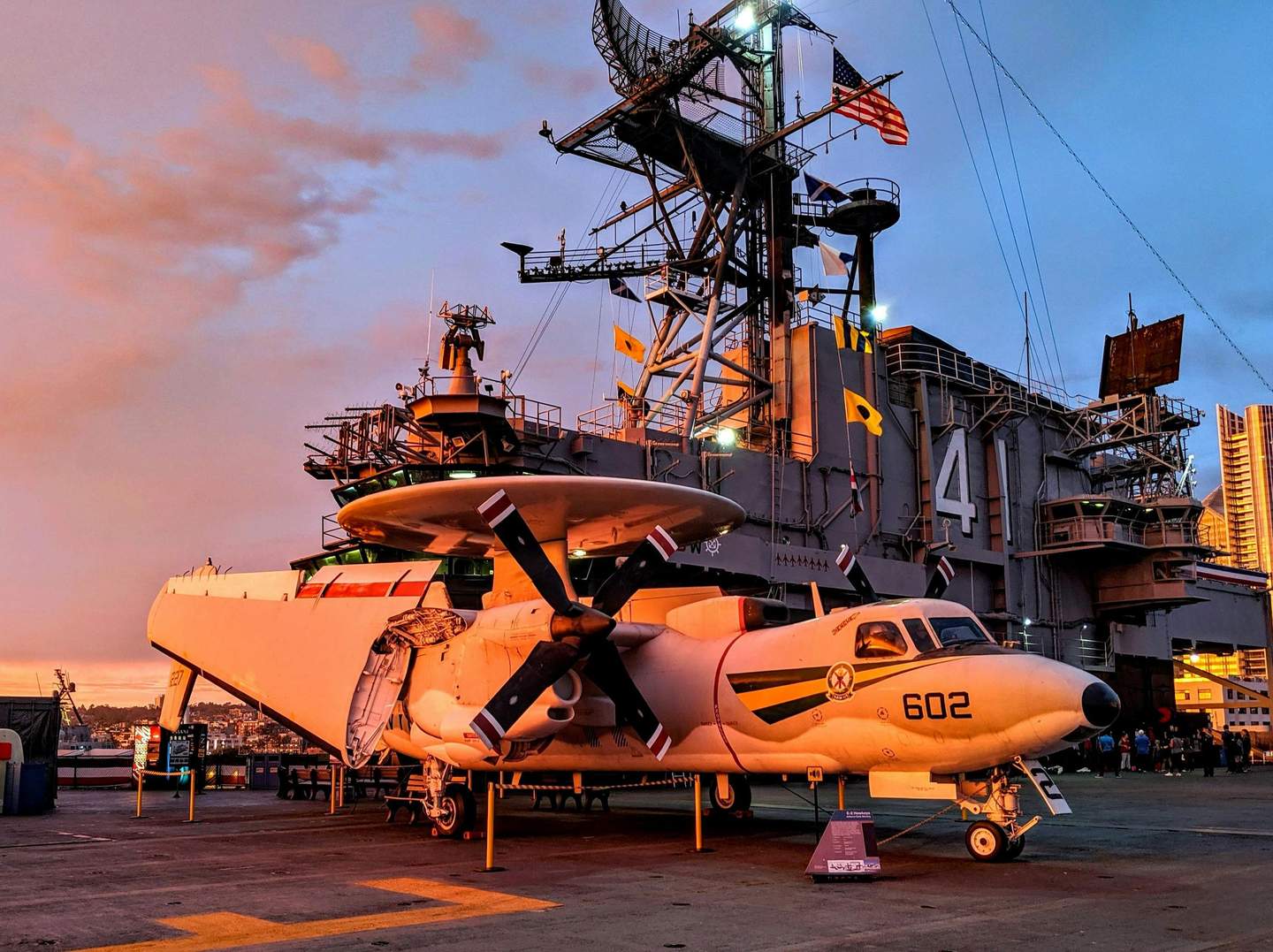 A white propeller aircraft marked "602" positioned on the deck of a naval ship at sunset, with an American flag flying.