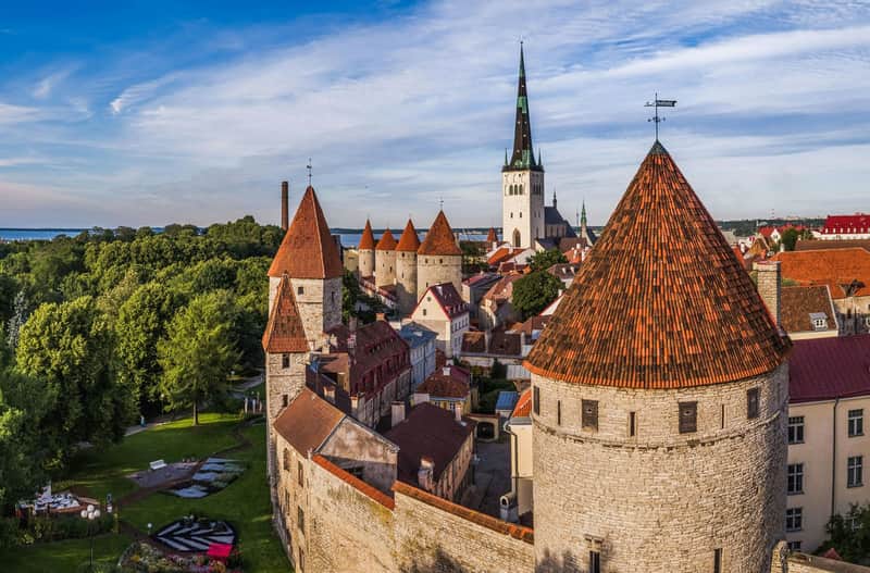 Aerial view of a historic town with stone walls, red-roofed towers, and a tall church spire surrounded by greenery.