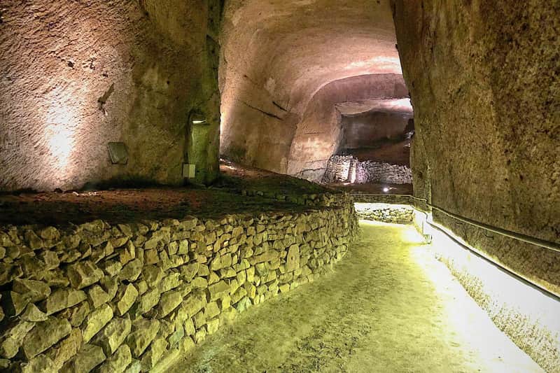 A stone-lined tunnel with illuminated pathways and arching, rough-hewn ceilings.