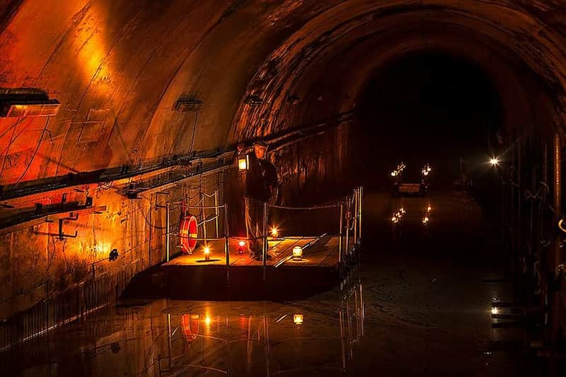 A person holding a lantern stands on a small, illuminated platform inside a dark, arched tunnel reflected in water.