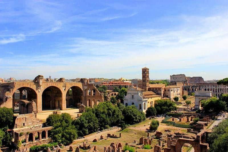 Ancient ruins and buildings under a partly cloudy blue sky with a mix of greenery and stone structures in the foreground.