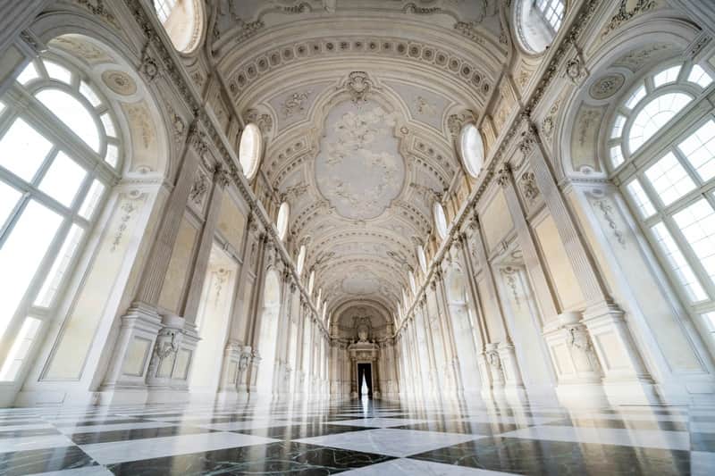 A grand hall with ornate vaulted ceilings, tall windows, and a checkered marble floor, leading to an open door in the distance.