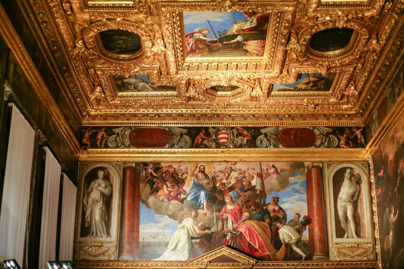 Combo do Palácio Ducal e da Basílica de São Marcos (Veneza) Sala ornamentada com um teto dourado e intrincadamente decorado e um grande mural que representa uma cena bíblica com várias figuras.