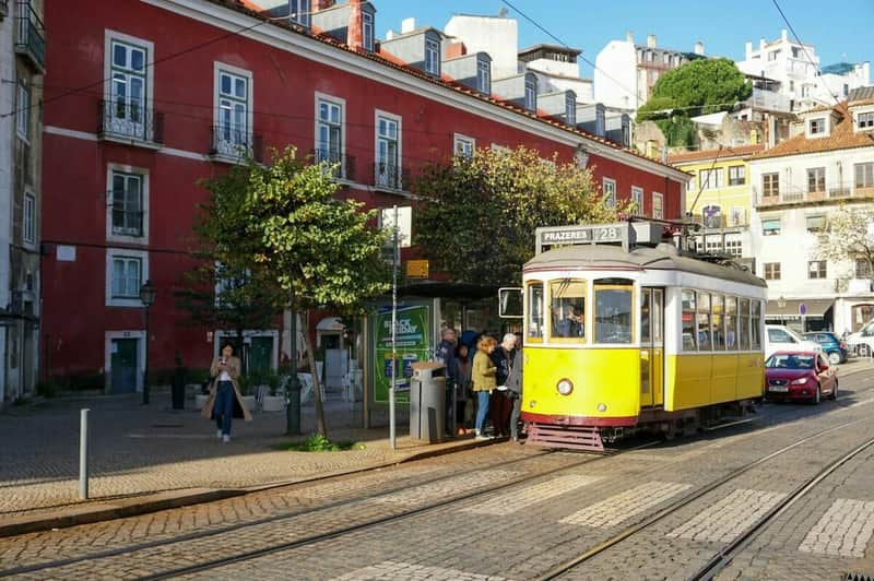 A yellow tram with passengers stopped in front of a red building, with trees and people walking nearby.