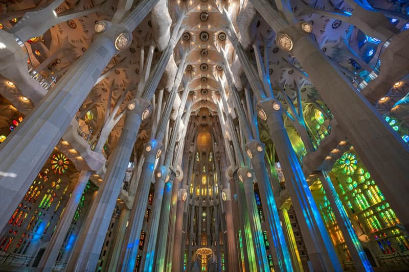 Barcelona Essentials Pass + Transportzugang (Barcelona) Das Innere einer Kathedrale mit hohen, verschnörkelten Säulen und bunten Glasfenstern, die vielfarbiges Licht auf die Wände werfen.