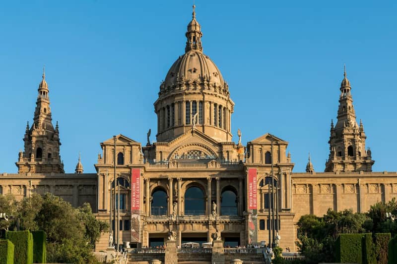 Barcelona Museumspass – Articket: Eintritt ohne Anstehen – Fundació Joan Miró und 4 weiteren Attraktionen Großes architektonisches Gebäude mit einer zentralen Kuppel und zwei Türmen, mit Bannern an der Fassade und einer Treppe im Vordergrund.