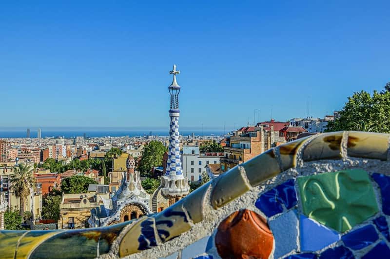 Barcelona: Park Güell + Seilbahn + Hop-on Hop-off Bus – Seilbahn Barcelona (Barcelona) Erhöhte Ansicht einer Stadtlandschaft mit einem hohen, blau-weiß gemusterten Kirchturm und buntem Mosaik im Vordergrund unter einem klaren blauen Himmel.