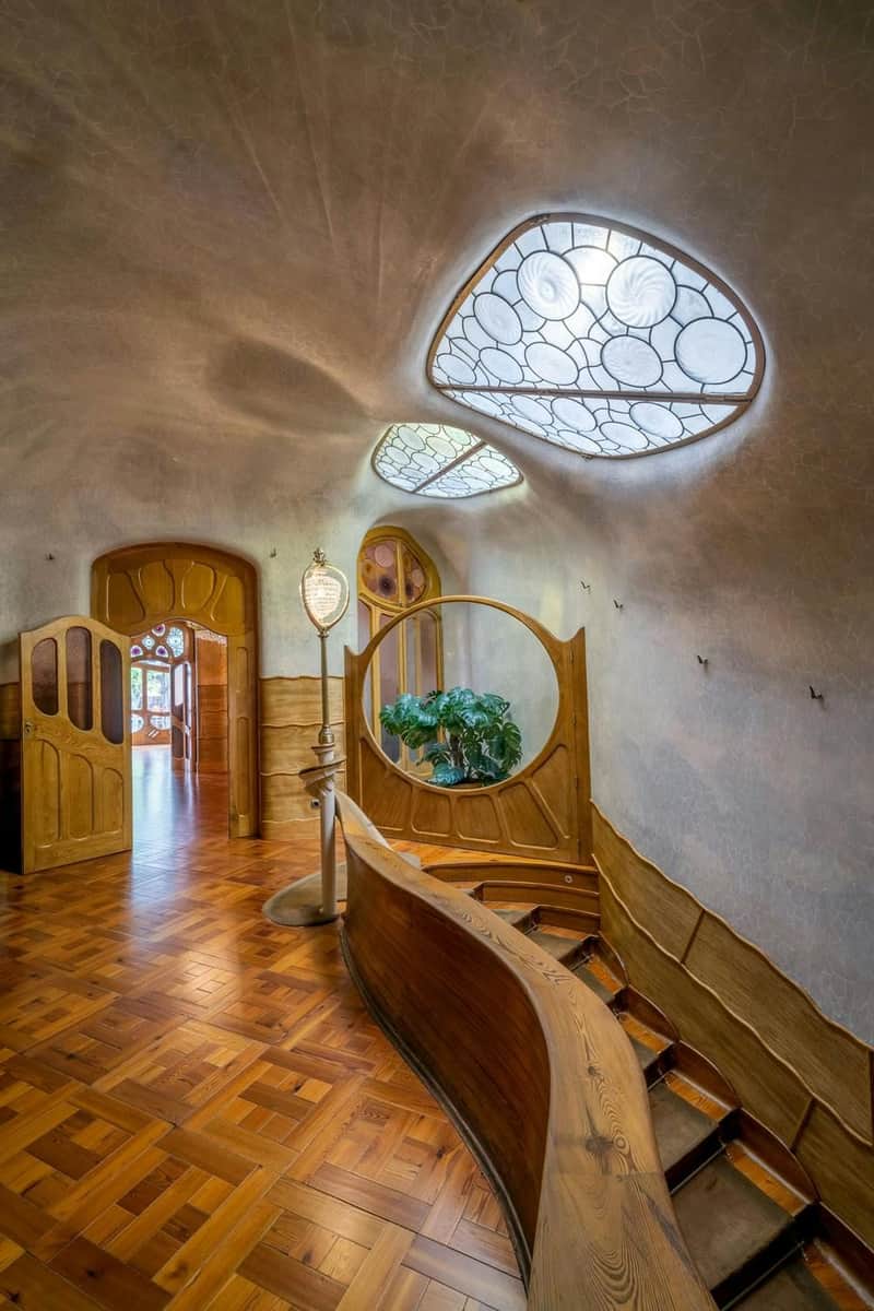 Casa Batlló + Hop-on Hop-off Bus Barcelona – Casa Batlló (Barcelona) Ein Flur mit verschnörkelten Holztüren, eine geschwungene Treppe, eine ausgestellte Pflanze und einzigartige geometrische Oberlichter an der Decke.