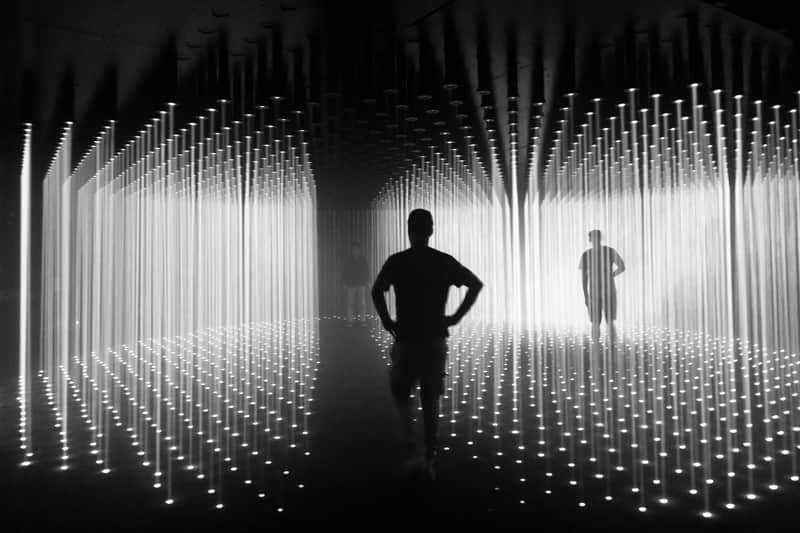 Silhouettes of people walking through a room with vertical light beams from ceiling to floor.