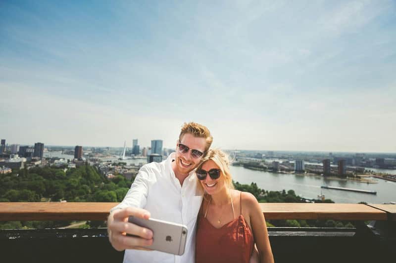 A couple wearing sunglasses takes a selfie on a sunny day with a cityscape and river in the background.