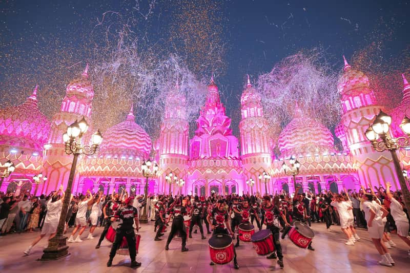 A vibrant celebration with performers in costumes playing drums in front of an illuminated, ornate building at night.