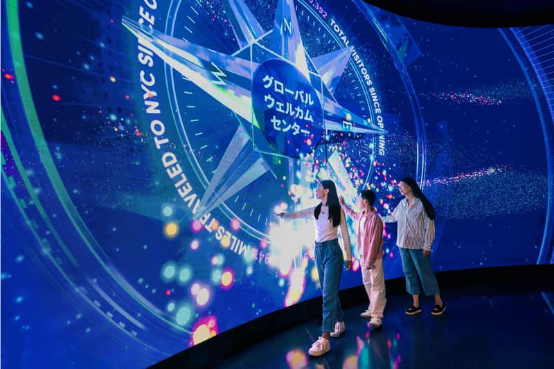 NYC Observation Decks Pass – Edge NYC and 3 other attractions Three people interact with a large digital display featuring colorful graphics and text in multiple languages, including Japanese.