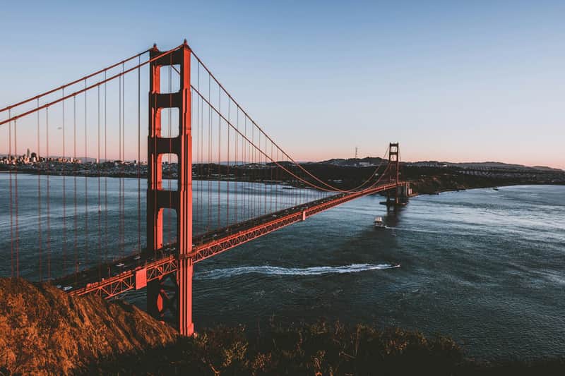 Red suspension bridge spans across a body of water at sunset, with a city skyline in the background. Boat leaves a wake in the water.