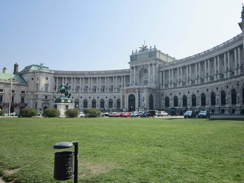 Hofburg Palace City Card (Vienna) Grand building with curved facade, statue of rider on horse in front, cars parked along the street, green lawn and trash can.