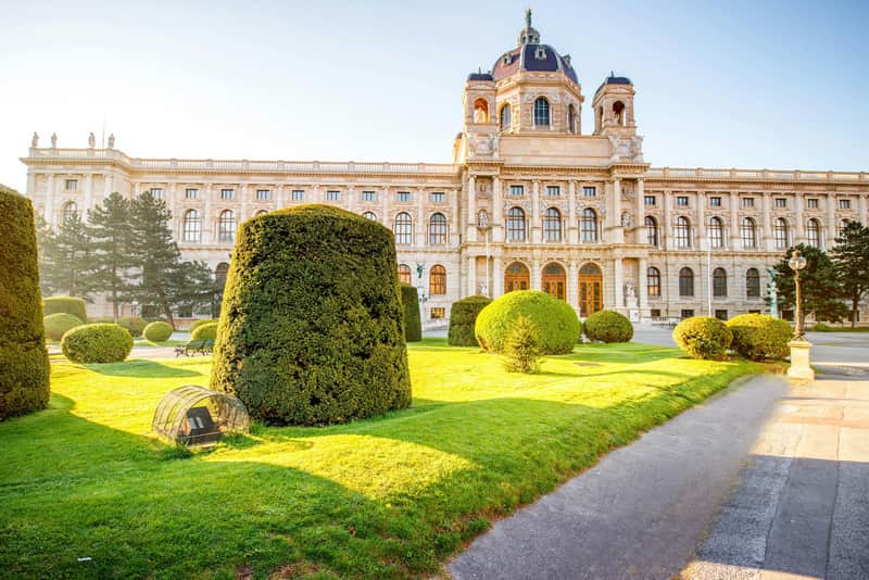 Kunsthistorisches Museum City Card (Vienna) A large, historic building with arched windows, a dome, and neatly trimmed hedges in a sunlit garden area.