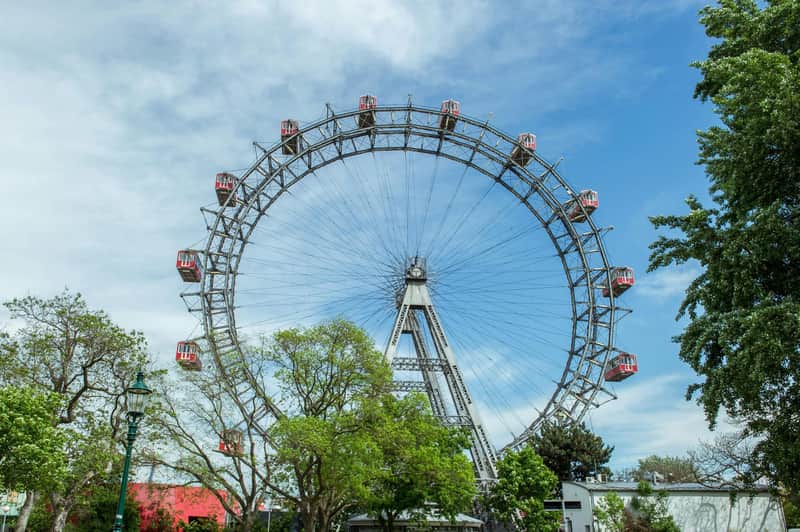 Wiener Riesenrad City Card (Vienna) A large Ferris wheel with red gondolas, surrounded by trees, under a partly cloudy blue sky.