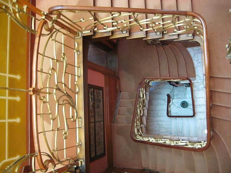 Looking down a spiral staircase with ornate golden railings, brown carpeted stairs, and landing areas with a green vase visible below.