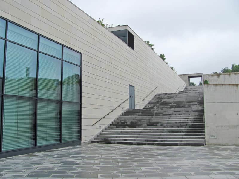 Modern building with large windows on the left and wide stone steps leading up to the right, surrounded by gray paving stones.