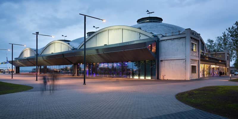 A modern building with large windows and industrial design features, under an evening sky with streetlights and silhouettes of people.