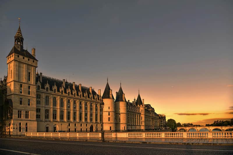 Historic building with tower and turrets along a river at sunset, with a bridge in the background and clear sky.