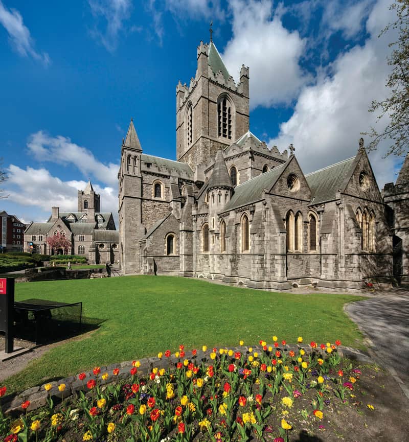 Gothic-style stone church with a tower, surrounded by green lawn and colorful tulip garden under a blue sky with clouds.
