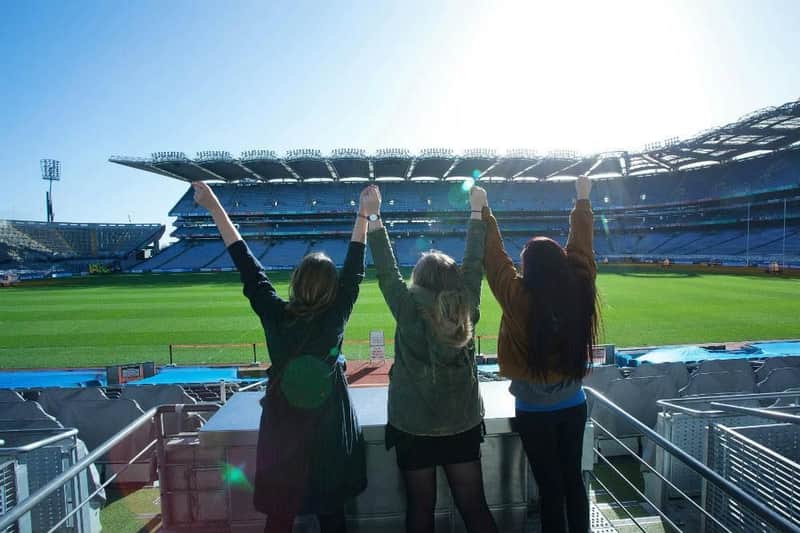 Three individuals with raised arms stand facing a large, empty stadium under bright sunlight.