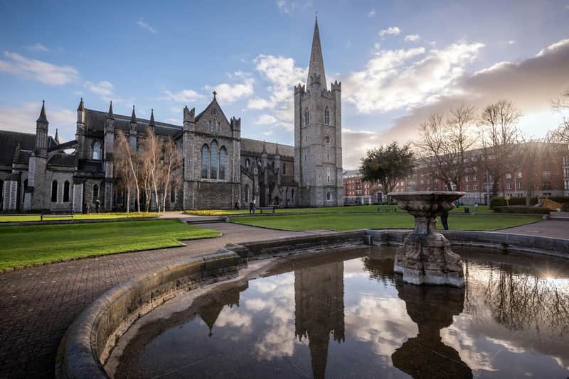 A historic stone cathedral with a tall spire beside a reflective fountain, surrounded by green lawns and a few people under a blue sky.