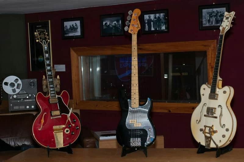 Three guitars on stands in a music studio, along with a reel-to-reel tape recorder. Framed photos hang on a burgundy wall.