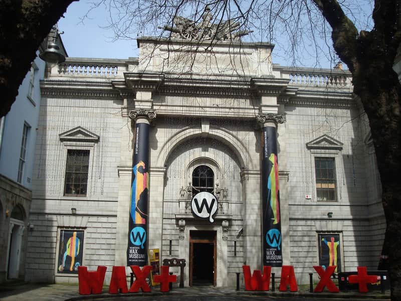 Front facade of the Wax Museum building with a stone exterior, large pillars, banners, and a "WAX+" sign.