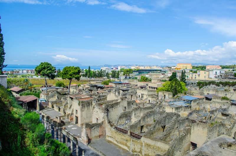 Ancient ruins with stone structures and columns, surrounded by greenery, with a town and sea in the background under a blue sky.