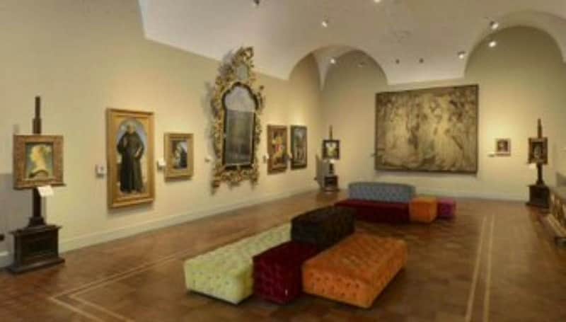 An art gallery room with paintings on the walls and multi-colored tufted benches in the center.