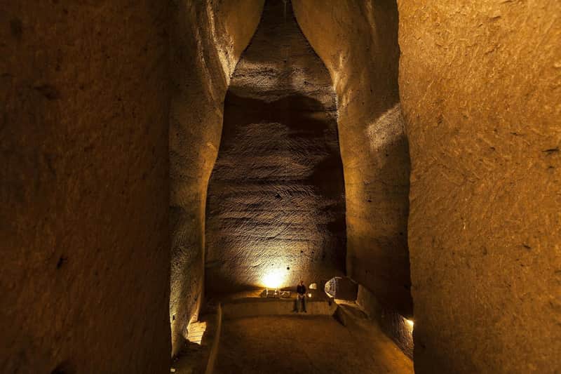 A person stands in a massive, dimly lit underground cavern with tall, textured walls and a spotlight illuminating the area.