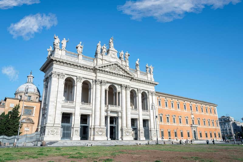Basilica di San Giovanni in Laterano City Card (Rome) Historic church facade with statues, arched windows, and columns under a clear blue sky, adjacent to a dome and orange building.