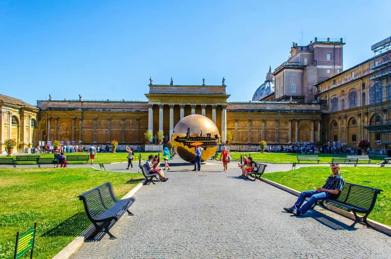 Vatican Museums City Card (Rome) Tourists relax on benches in a courtyard with a large bronze sculpture and historic building under a clear blue sky.