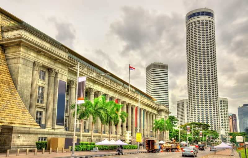 Vista de una calle con un edificio histórico con columnas, un asta de bandera y palmeras, con edificios modernos de gran altura al fondo.
