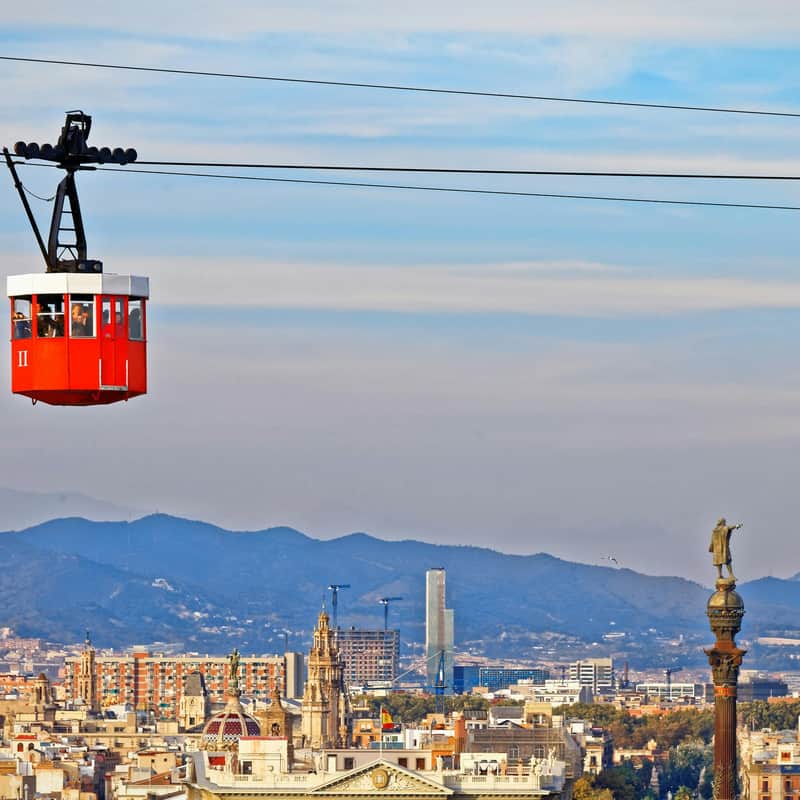 Targeta de la ciutat per a Telefèric de Barcelona (Telefèric del Port) (Barcelona) Un telefèric vermell travessa el cel amb un paisatge urbà i muntanyes al fons sota un cel ennuvolat.