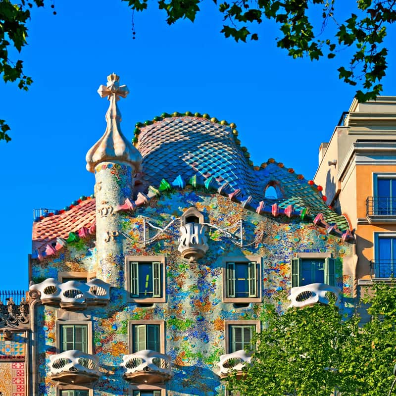 Targeta de la ciutat per a Casa Batlló (Barcelona) Un edifici de colors cobert de mosaics amb balcons corbats, persianes verdes i una torreta en forma de con, emmarcada per branques d'arbres.
