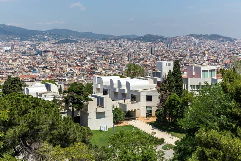 Fundació Joan Miró Bykort & Pas (Barcelona) Med udsigt over en tæt bygget by med hvide, moderne bygninger og omkringliggende grønne områder i forgrunden. Fjerntliggende bjerge er synlige.