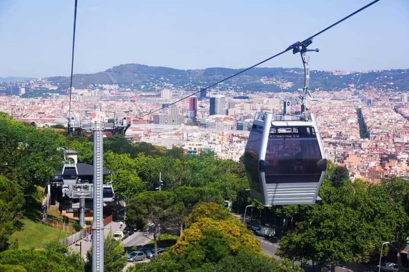 Κάρτα Πόλης για Τελεφερίκ Montjuïc (Μοντζουίκ) (Βαρκελώνη) Τελεφερίκ που κινούνται πάνω από ένα καταπράσινο τοπίο με πυκνό αστικό τοπίο και βουνά στο παρασκήνιο.