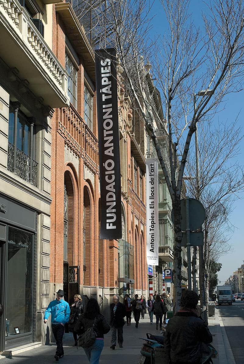 Museu Tàpies City Cards & Pässe (Barcelona) Die Menschen gehen am Gebäude der Fundació Antoni Tàpies entlang, das an einer belebten, von Bäumen gesäumten Straße mit zahlreichen Ladenfronten liegt.