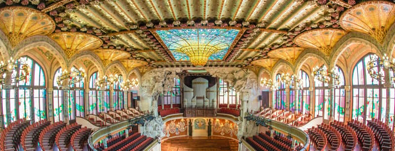 Targeta de la ciutat per a Palau de la Música Catalana (Barcelona) Sala de concerts ornamentada amb vitralls en arc, dissenys de sostre intrincats, candelabros i files de seients vermells.