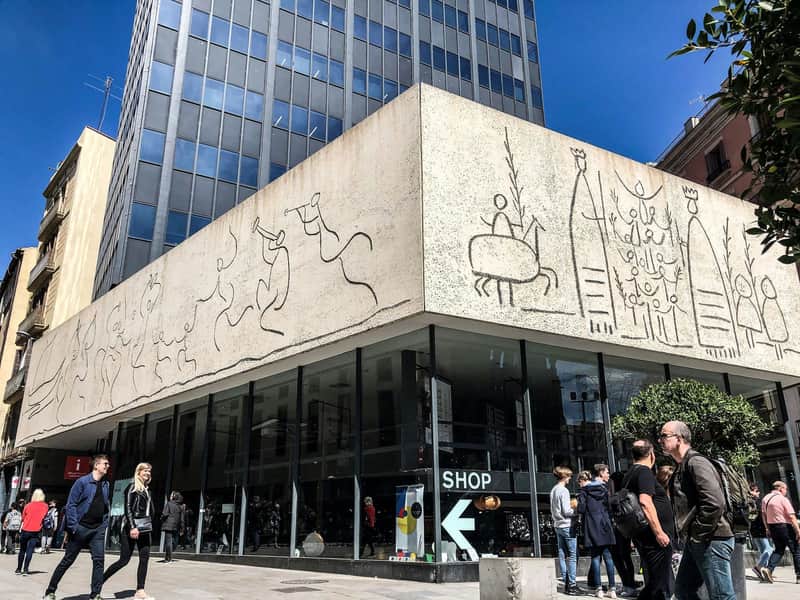 Targetes de la ciutat per a Museu Picasso (Barcelona) Un edifici modern amb dibuixos de línies a la façana, gent passant i una botiga amb vidre a peu de carrer.