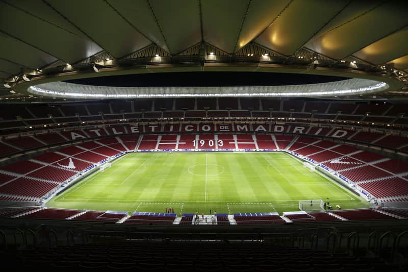 Atlético de Madrid City Card (Madrid) Empty football stadium with red seats, green pitch, and "Atletico de Madrid" written on the stands, under a roof with lights.